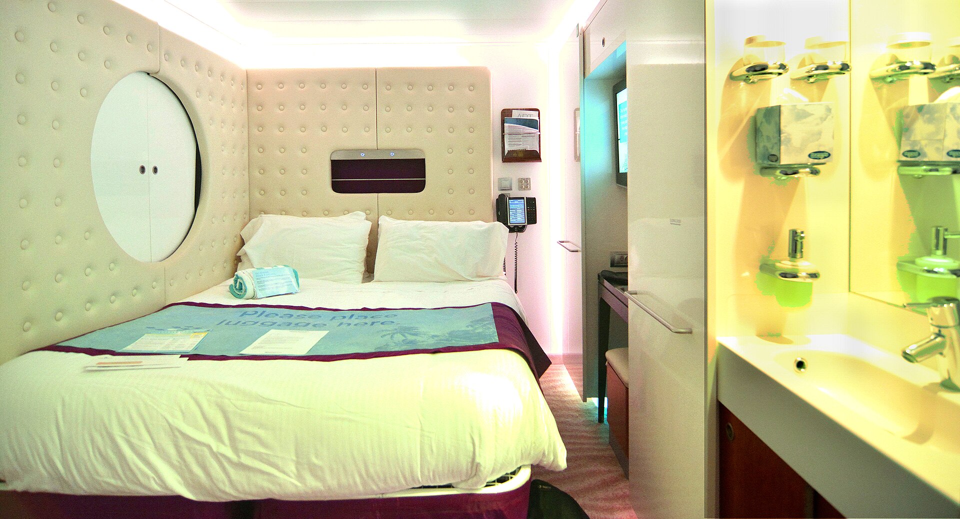 A compact solo studio cabin on Norwegian Epic with a double bed, padded headboard wall, round porthole-style cupboard and a glowing washbasin