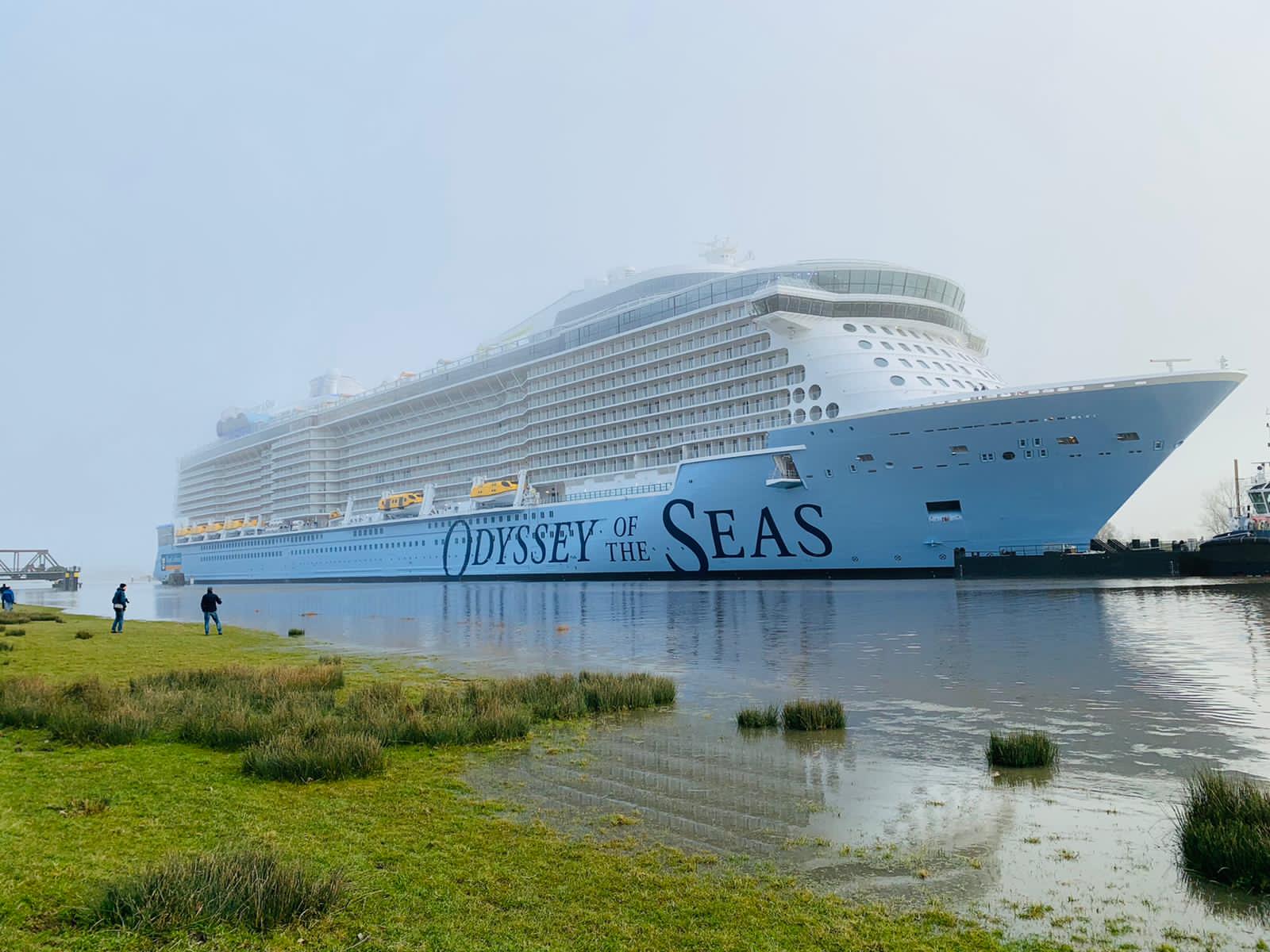 Royal Caribbean's Odyssey of the Seas with her blue hull and name in large letters, seen from a grassy riverbank