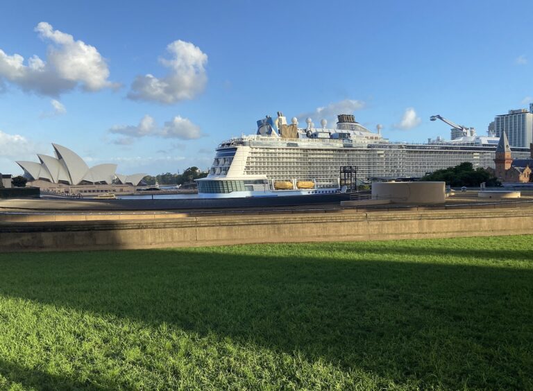 Royal Caribbean's Ovation of the Seas docked in Sydney under a bright blue sky, with the Opera House and a green lawn in the foreground