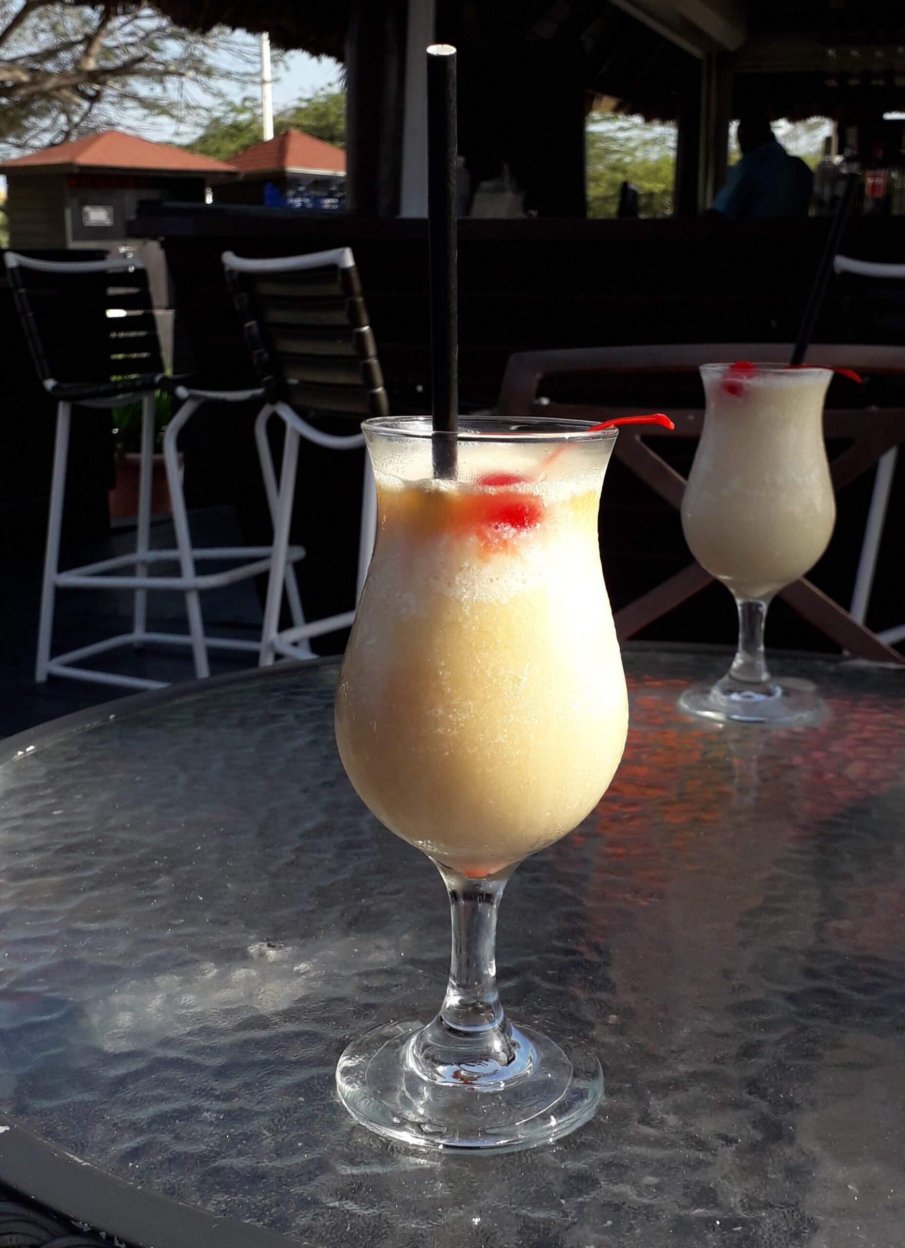 Two frozen pina colada cocktails with cherries on a glass table at an outdoor Caribbean bar
