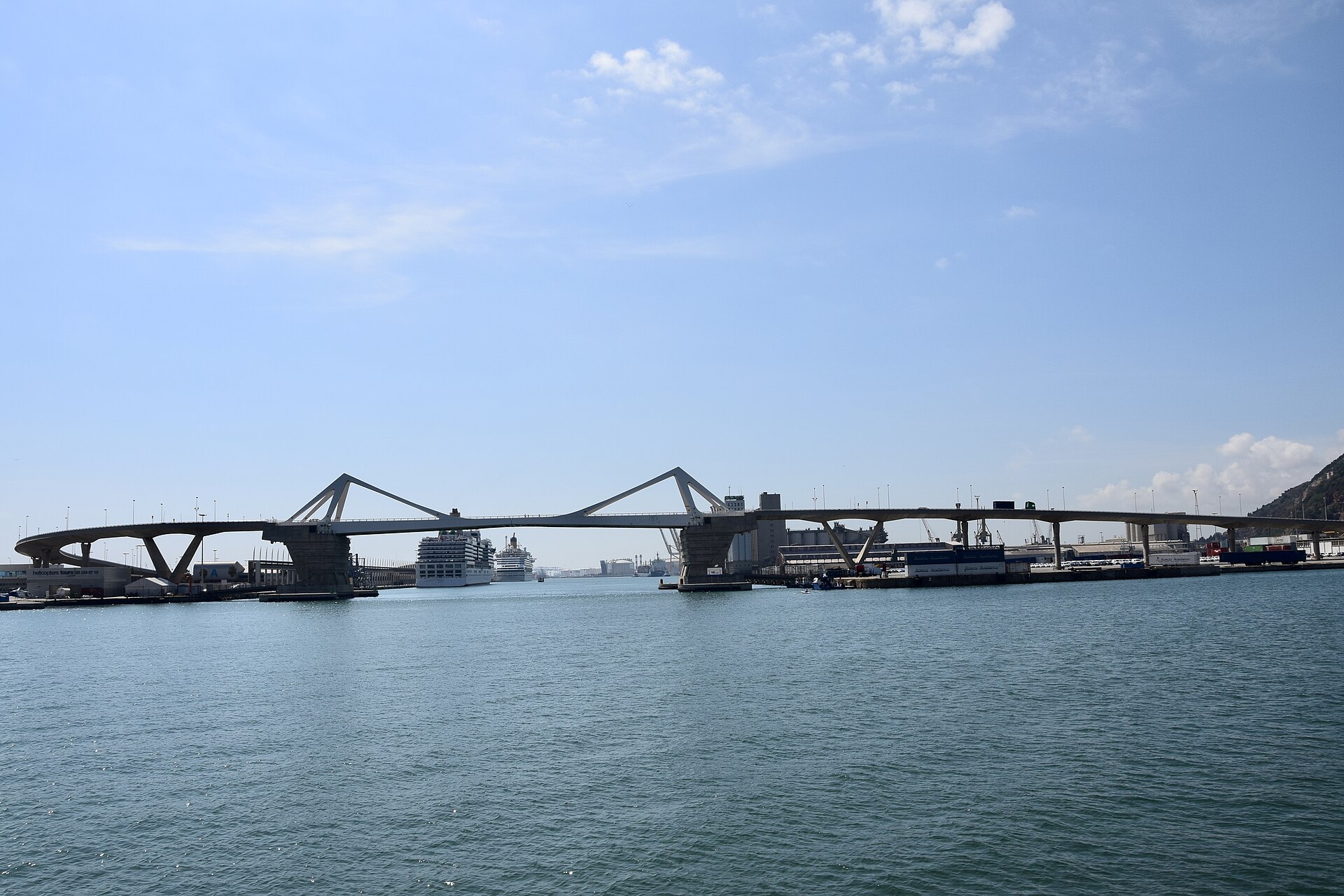 The Porta d'Europa bascule bridge in the Port of Barcelona under a blue sky, with cruise ships beyond it
