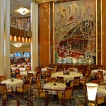 The Britannia Restaurant on Queen Mary 2, with round tables in white linen, honey-coloured chairs and a huge Art Deco mural of a liner in New York