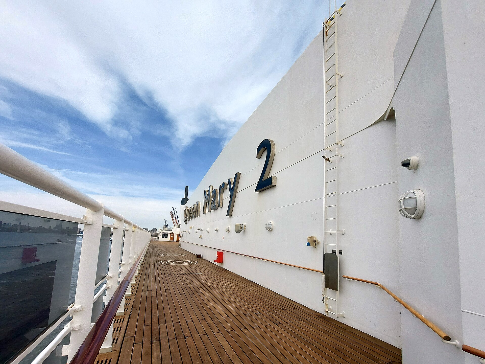 Teak top deck of Cunard's Queen Mary 2 with the ship's name on a white wall under a blue sky in Brooklyn