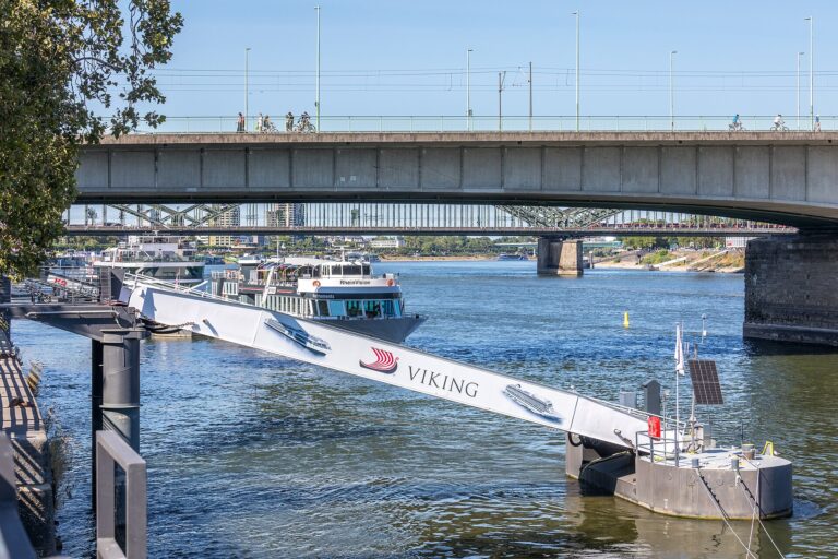 A Viking river cruise gangway slopes steeply down to a pontoon on the low Rhine in Cologne, with the Deutz Bridge behind