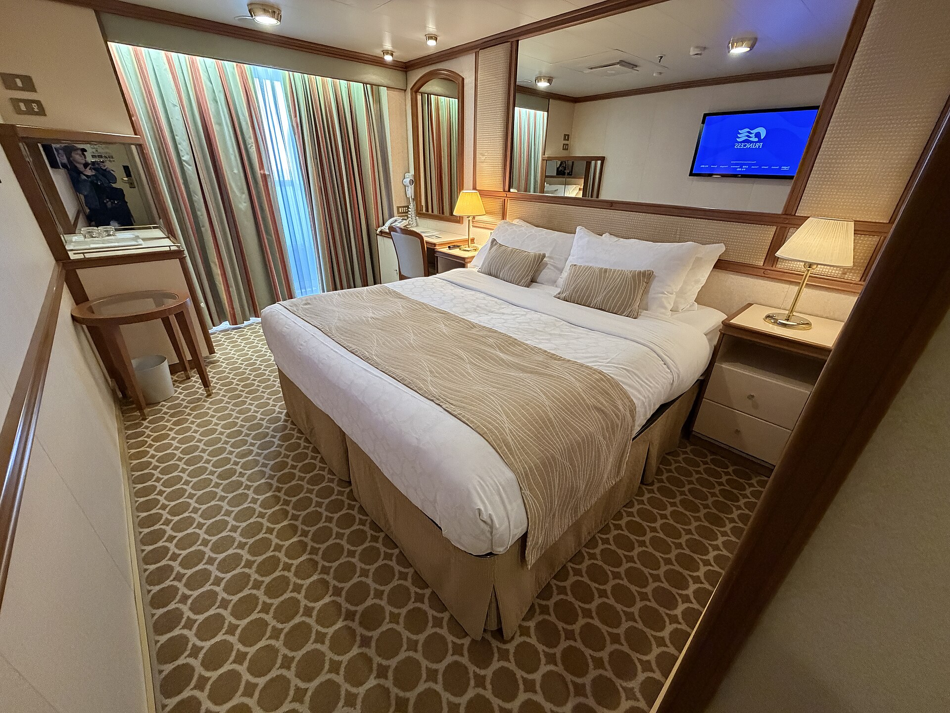 A balcony cabin on Ruby Princess with a queen bed, bedside lamps, a desk and a TV, and curtains half open to the balcony door