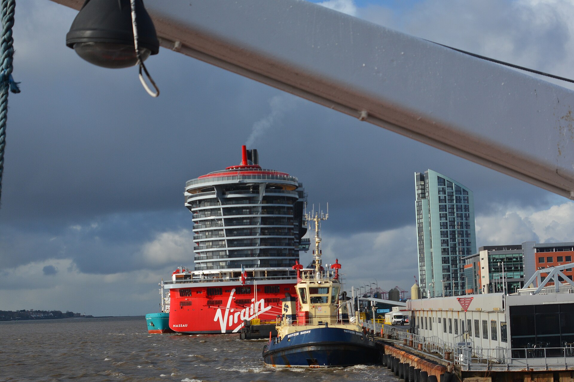 The red stern of Virgin Voyages' Scarlet Lady with the Virgin logo, seen past a harbour tug