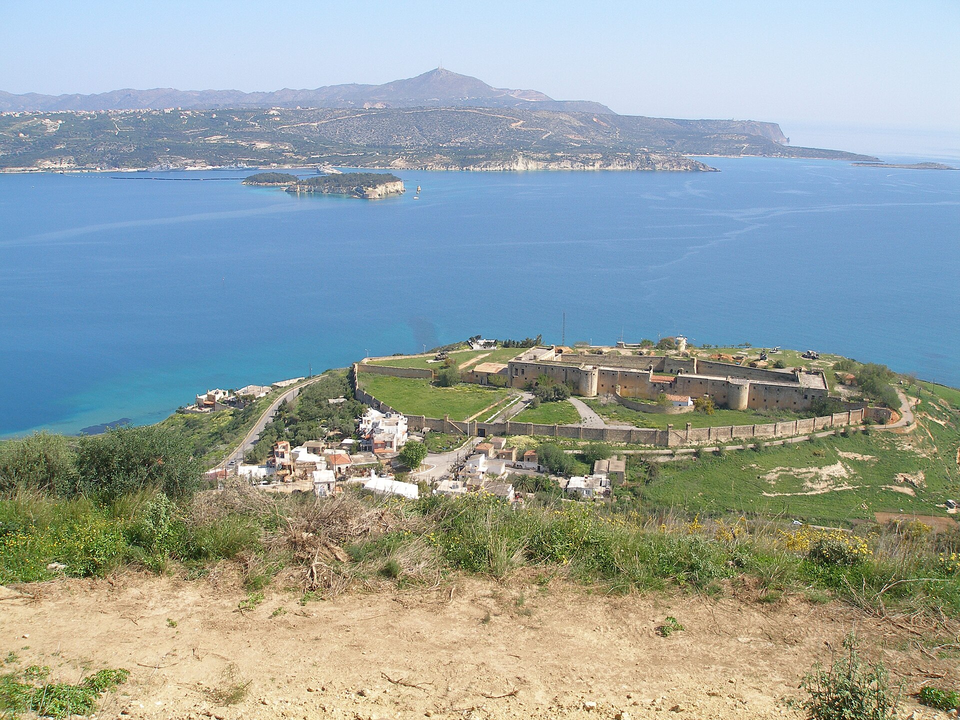 The entrance to Souda Bay in Crete, a long blue inlet ringed by hills, with an old fortress in the foreground