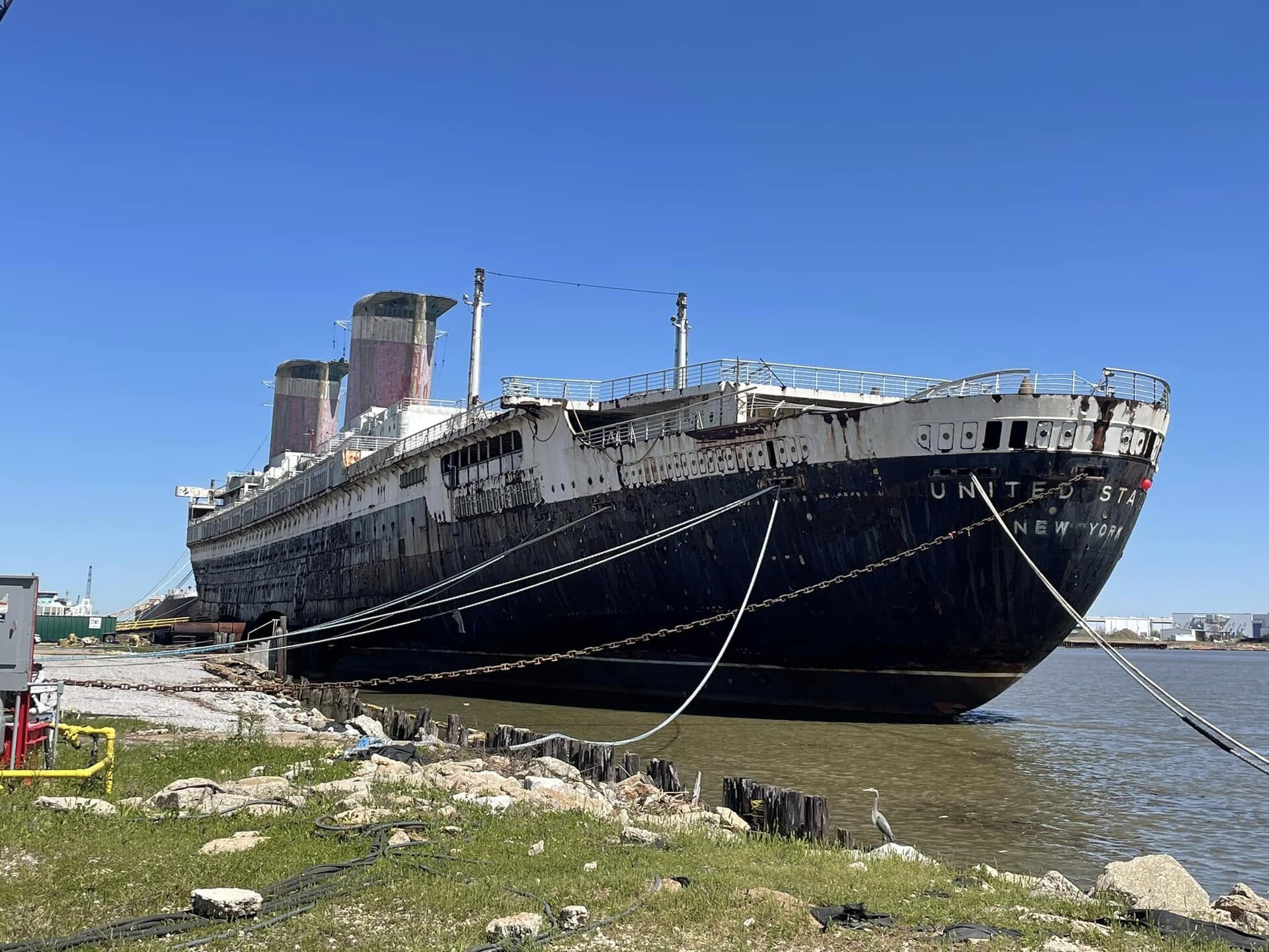 The SS United States docked in Mobile, Alabama, under a clear blue sky with a heron on the shore
