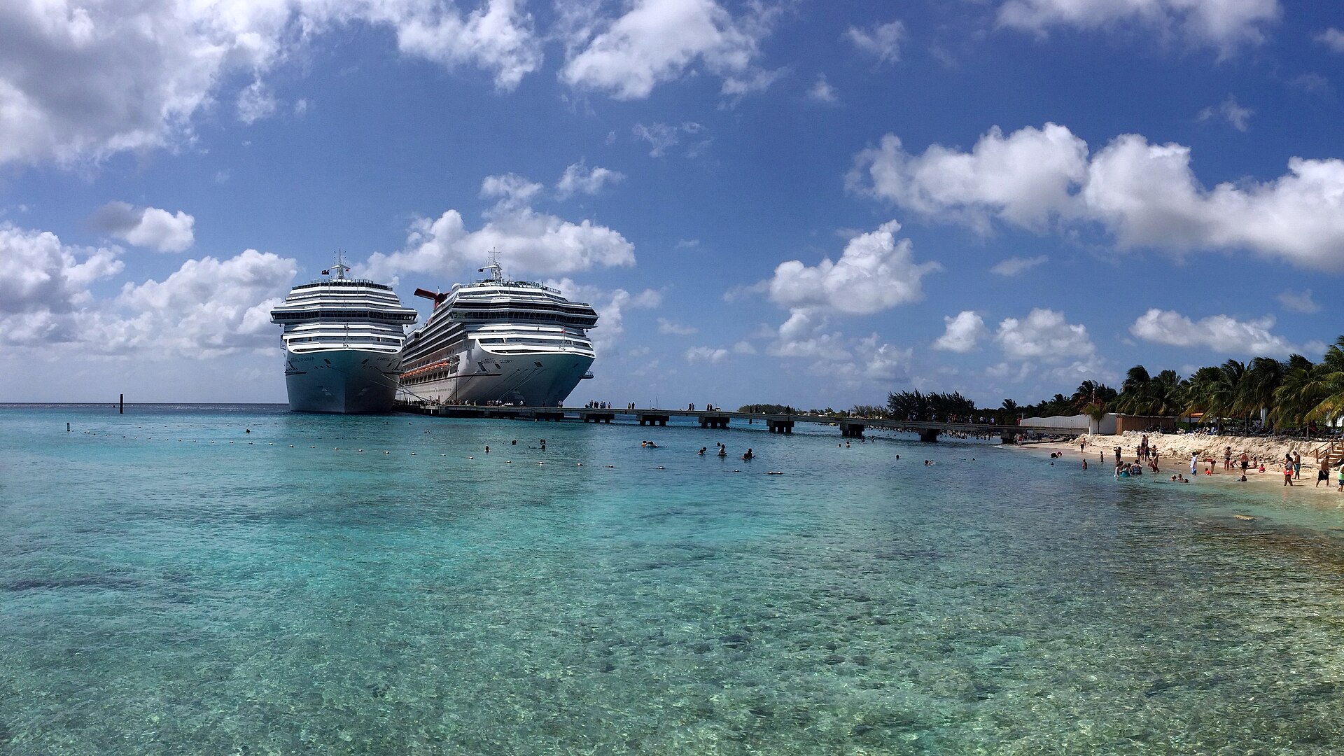 Two Carnival cruise ships docked at Grand Turk beside clear turquoise water and a palm-lined beach
