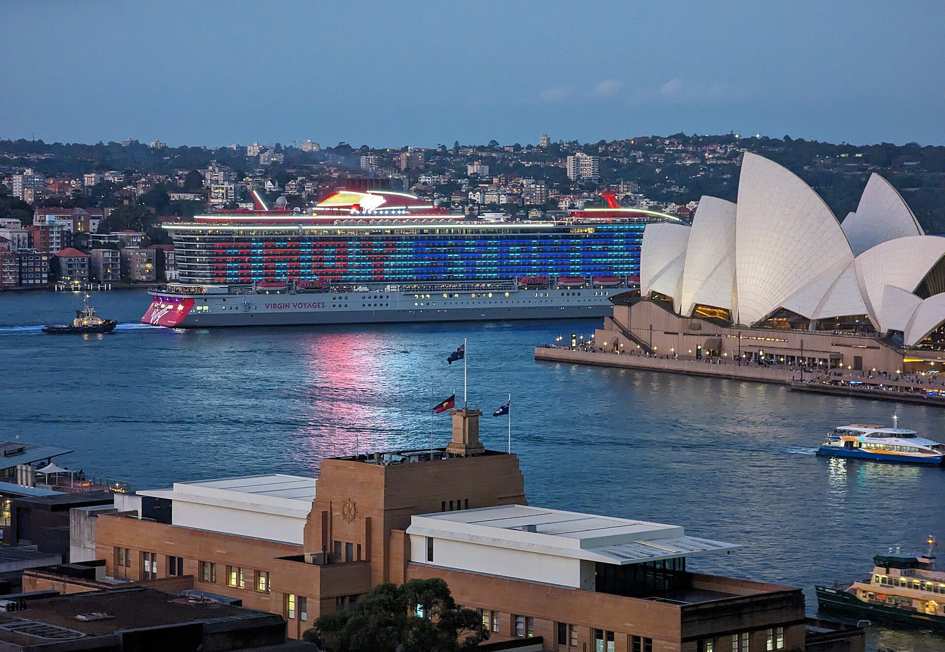 Virgin Voyages Resilient Lady, lit with AHOY SYDNEY on her side, sailing past the Sydney Opera House at dusk